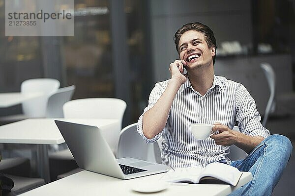 gutaussehender kaukasischer junger Mann mit Telefon  der am Laptop arbeitet und lächelt  während er einen Kaffee im Café genießt