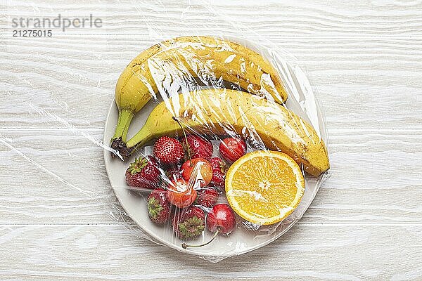 Assorted verschiedene Früchte und Beeren auf weißem Teller in Kunststoff eingewickelt klar transparente Folie von oben auf weißem Holz rustikalen Tisch Hintergrund  Lebensmittel Fotografie