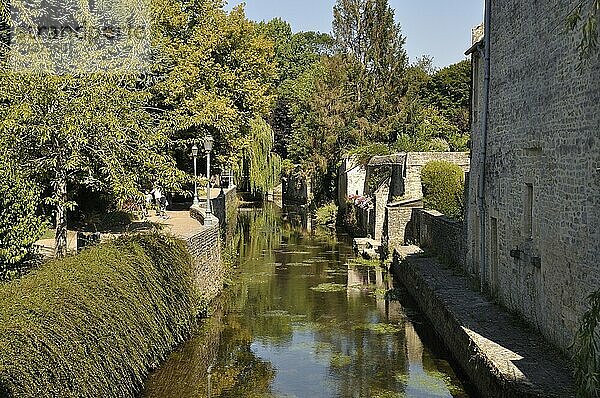 Ein ruhiger Fluss  flankiert von Steinmauern und üppigem Grün  mit Spiegelungen von Bäumen und einem Gebäude  die eine heitere Atmosphäre schaffen: Der Fluss Aure in Bayeux