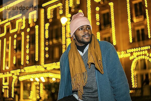 Afrikanischer Mann  der in der Weihnachtsnacht im Freien sitzt und auf die Lichterketten blickt  die eine Fassade schmücken