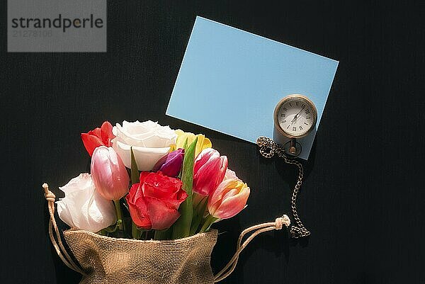 Bunter Strauß verschiedener Blumen in einem Jutebeutel  eine alte Taschenuhr und eine leere blaue Nachrichtenkarte  auf schwarzem Hintergrund