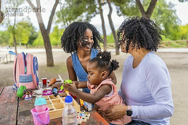 Glückliches afrikanisches Mädchen  das mit Spielzeug spielt und mit seiner Mutter und Großmutter in einem Park sitzt