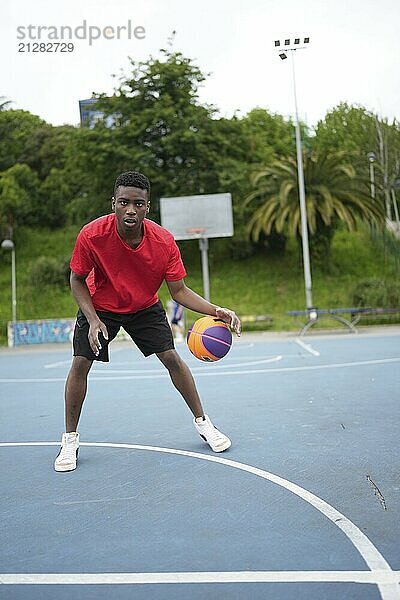 Vertikales Foto eines afroamerikanischen Basketballspielers in voller Länge  der allein auf einem Platz im Freien dribbelt