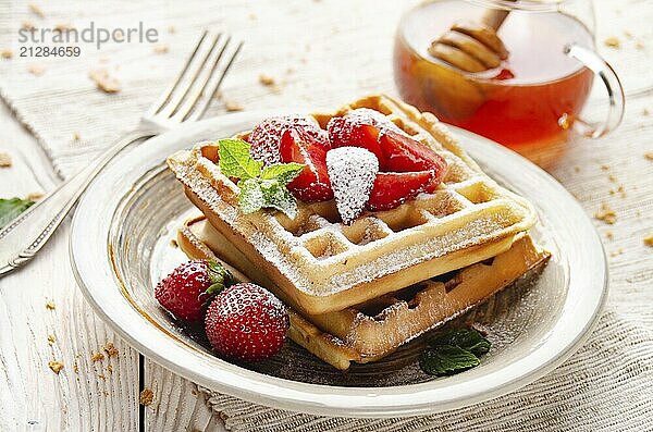 Belgische Waffeln  serviert mit Erdbeeren und Minzblättern  bestäubt mit Puderzucker auf einem weißen Holztisch mit Sirup daneben