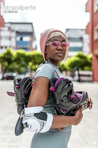 Vertikales Foto einer selbstbewussten urbanen afrikanischen Frau mit Inline Skates und Schutzausrüstung für die Ellbogen  die in der Stadt posiert