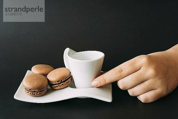 Frau  die eine Tasse Kaffee und Macarons vor schwarzem Hintergrund zu sich nimmt. Selektiver Fokus