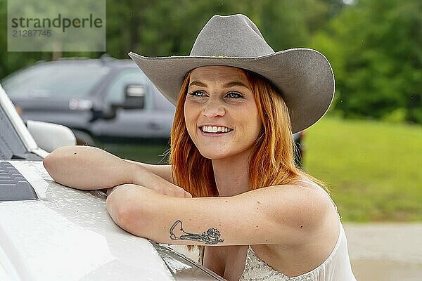 Ein schönes rothaariges Model posiert in einer ländlichen Umgebung in Cowgirl Kleidung vor ihrem Truck