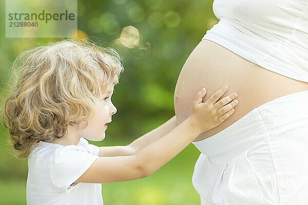 Glückliches Kind hält Bauch der schwangeren Frau vor grünem Frühlingshintergrund