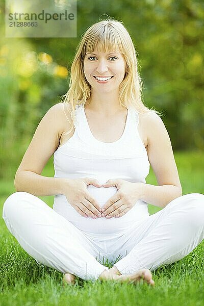 Glückliche schwangere Frau sitzt auf grünem Gras und hält die Hände auf den Bauch in Form von Herz