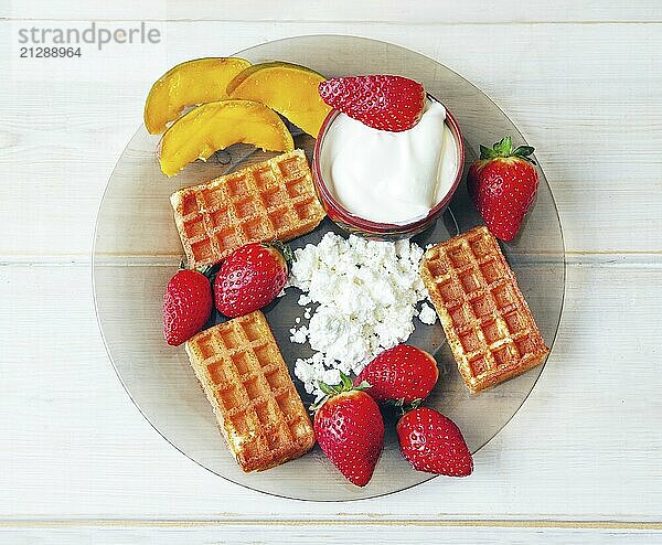 Saure Sahne und Hüttenkäse und Waffeln und Erdbeeren und Mangostücke auf weißen Holzbrettern