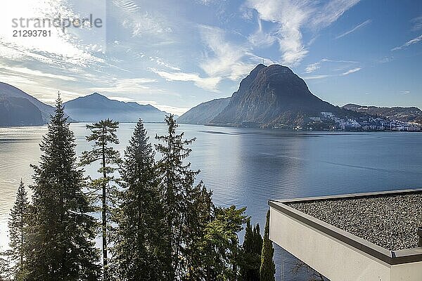 Sunny view of Lake Lugano and nice residential buildings on the shore (roofs of 2 apartment buildings) with a fantastic cedar and Mount San Salvatore in the distance and a bridge over the lake. On the roof of the building  white pebbles