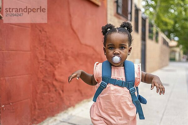 Cute baby african girl running along a street using pacifier