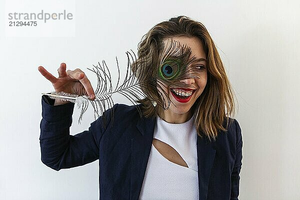 A close up and front portrait of a flirtatious brown haired woman standing against a white background  holding a peafowl tail over one eye and laughing