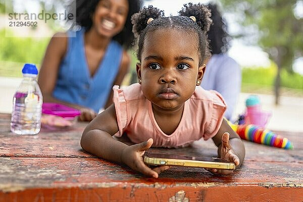 Close-up of a cute African baby girl using mobile phone in the park while her mother and granny talking on the background