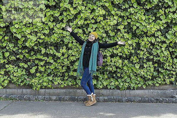 Oyful Latino woman in her 30s with arms wide open celebrating life in front of a vibrant green plant wall