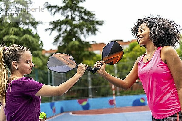 Team of two multi-ethnic women clashing rackets before playing pickelball in an outdoor court