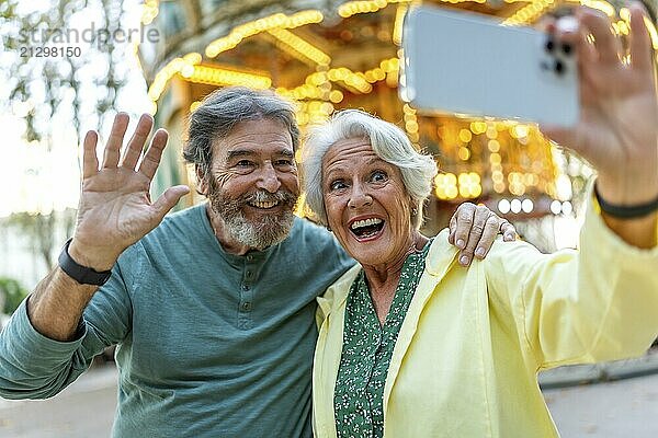 Senior couple waving at camera while taking selfie in the city next to carousel