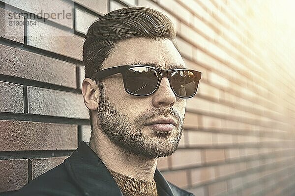 Portrait of stylish handsome young man with bristle standing outdoors and leaning on brick wall. Man wearing jacket  sweater and sunglasses