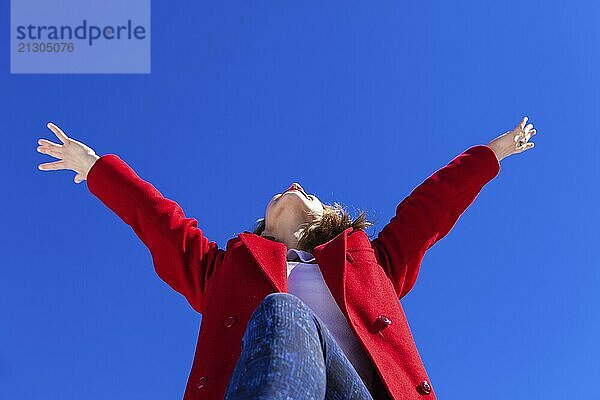 An elated Caucasian woman  wearing a red coat  has her arms in the air with joy on a bright winter's day against a blue sky  shot from a low angle perspective