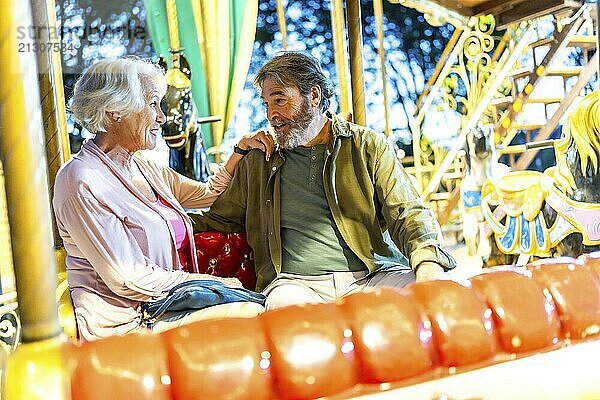 Senior modern caucasian playful couple sitting on carousel enjoying night together in the city