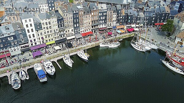 Fantastic drone point of view on Normandian ancient Honfleut town with harbor of sailboats and fishing boats  And outdoors cafes and people sittinf and walking (distant view. Therelandmark of old town with very small house ib right corner (for one family?) and more large building in centre part