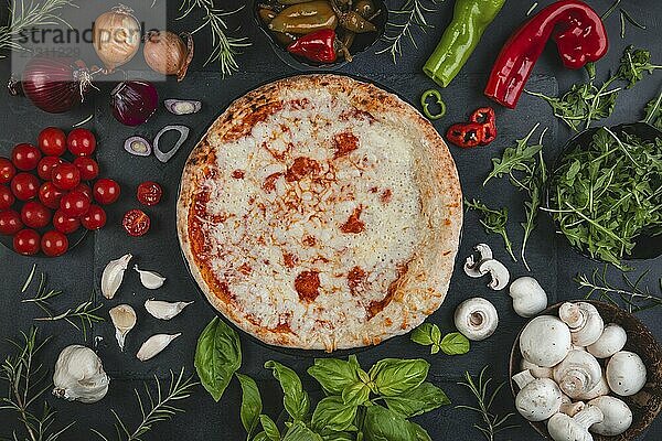 Pizza surrounded by fresh ingredients such as tomatoes  herbs  peppers  mushrooms and onions on a dark background