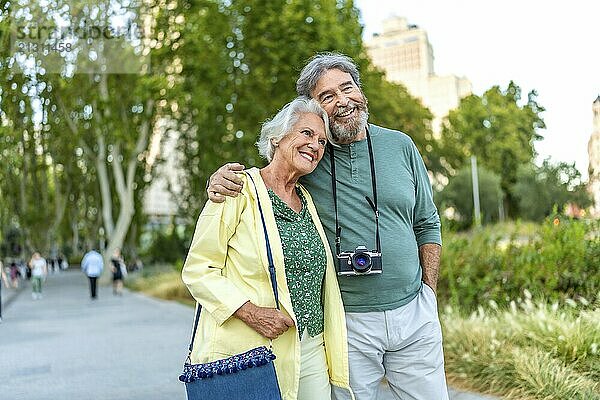 Cute senior lovers walking along an urban park carrying handbag and photographic camera