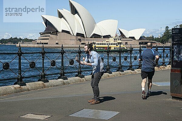 25.09.2019  Sydney  New South Wales  Australia  A man holds an ice cream in his hand and takes a photo with his mobile phone while a jogger runs past him and the famous Sydney Opera House can be seen in the background  Oceania