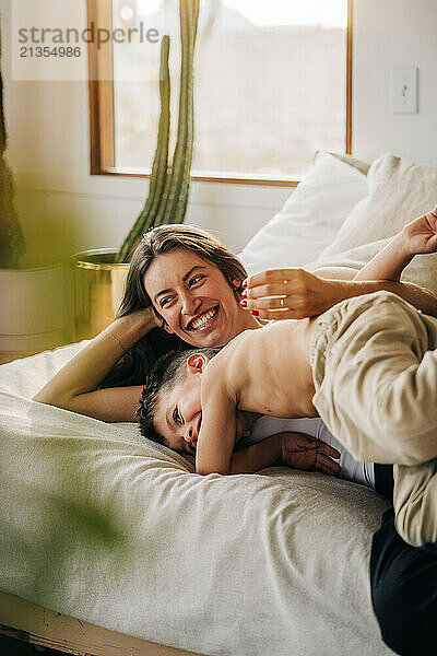 Mother and young son share a joyful cuddle on a cozy bed by the window