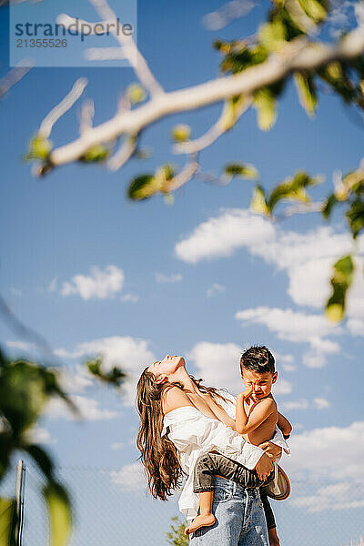 Woman joyfully holding young boy under blue sky with tree branches