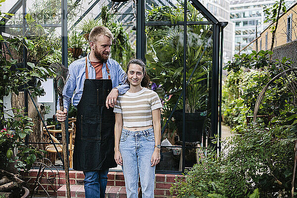 Man in apron standing with hand on shoulder of girlfriend outside greenhouse