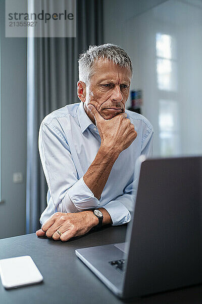 Senior businessman sitting with hand on chin and using laptop at desk in office