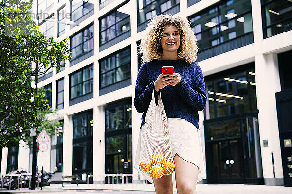 Happy curly haired woman standing with smart phone and bag of oranges near building