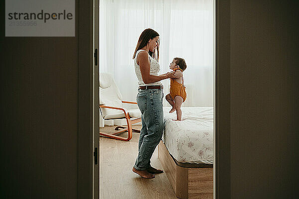 Woman playing with daughter standing on bed at home