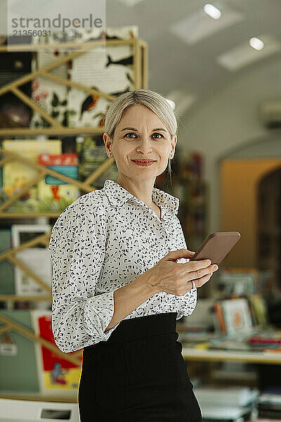 Confident businesswoman holding mobile phone in bookstore