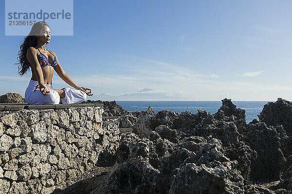 Yoga teacher sitting in lotus position on cliff of Mt Agung under blue sky