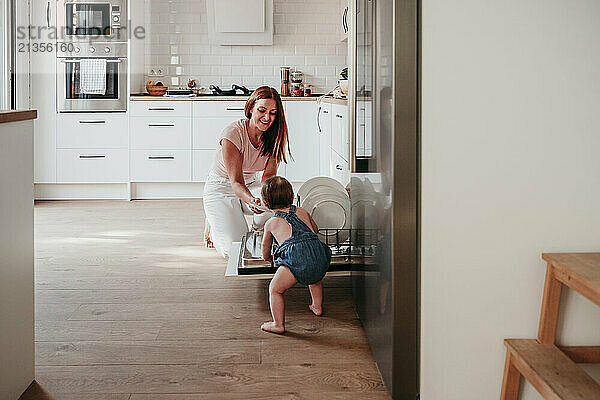 Baby girl standing near dishwasher with mother in kitchen at home