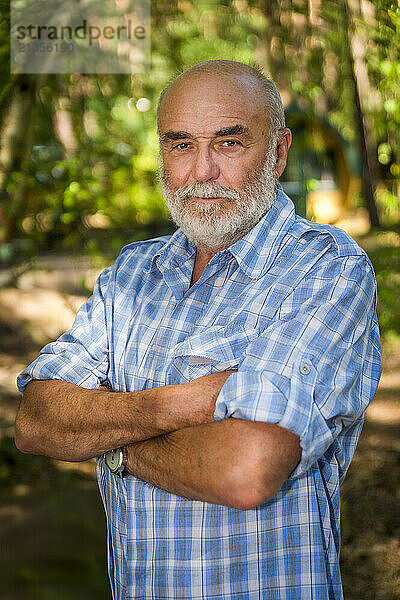 Smiling senior man standing with arms crossed in front of trees