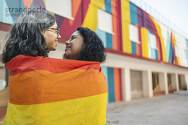 Lesbian couple wrapped in LGBTQIA flag standing in front of building