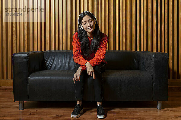 Smiling young woman listening to music through wireless headphones on leather sofa