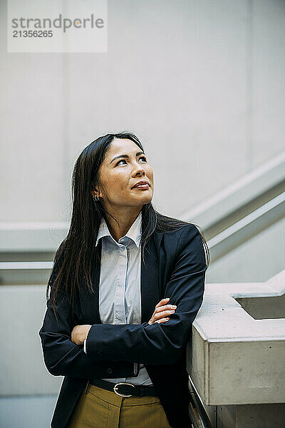 Businesswoman with arms crossed leaning on staircase railing looking up at office