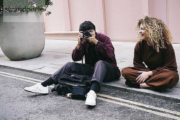 Happy woman sitting with friend photographing through camera on footpath