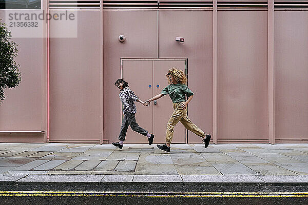 Happy lesbian couple holding hands and running in front of pink colored building