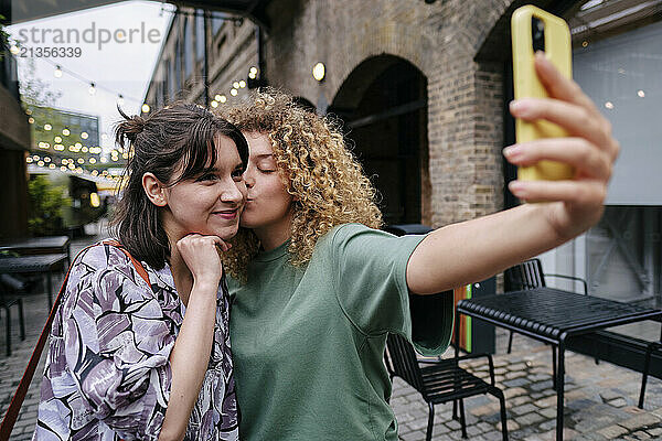 Woman kissing girlfriend and taking selfie through smart phone near building