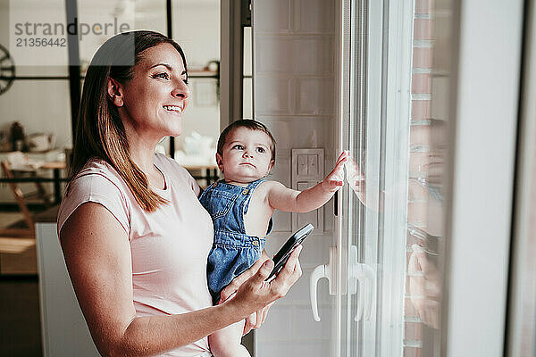 Smiling woman carrying daughter and holding smart phone standing near window at home