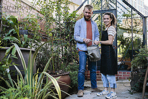 Couple holding watering can and standing outside greenhouse