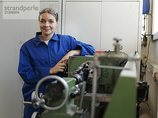 Smiling teenage trainee leaning on machine at workshop