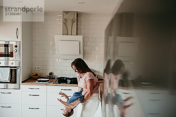 Cheerful mother playing with baby girl in kitchen at home