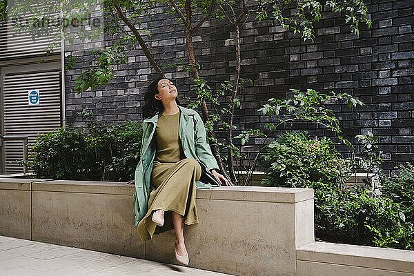 Woman sitting with eyes closed near plants in front of brick wall