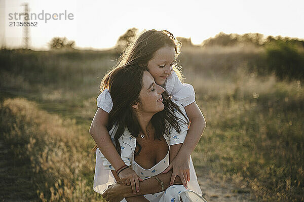 Smiling daughter hugging mother on field at sunset